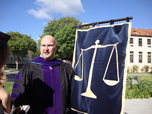 A male graduate wearing a black robe with purple accents holds a blue flag depicting scales.