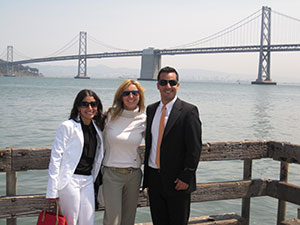 Three students--two female, one male--pose on a  wooden dock, in view of a long, gray bridge.