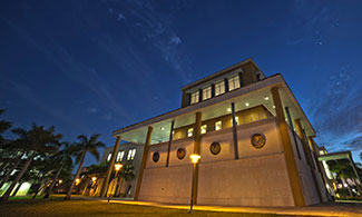 Angled view of a university building at night. Palm trees stand next to the building. A row of lit lampposts line the front of the building.