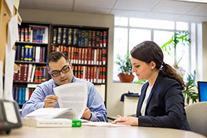 A man and a woman sit at a table indoors, each looking at papers. There is a large bookshelf behind them, filled with many sets of books. Green potted plants sit in a window.