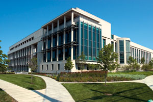 Law school exterior—a white building with a tall block of teal windows. Small deciduous trees are scattered among grass plots that lie in front of the school. The grass plots are divided by swooping paths.