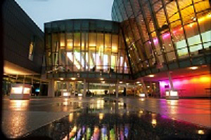 Colorful neon lights reflect off of the glassy university building and onto a wet, concrete walking path on a gray day.