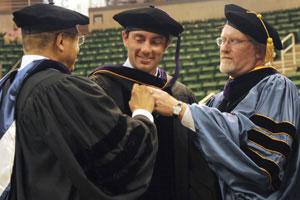 Two male professors and a male graduate huddle in the auditorium before the ceremony.