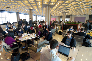 Students are crowded in a busy, well-lit study area. Some have food and cups of coffee beside their open laptops.