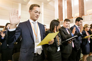 Students standing in rows raise their right hands while reading from sheets of paper.