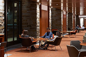 Four students converse at a table in a long room. Another group of students converses at the far end of the room.