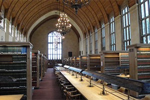 A long study table runs through the center of the law school library, toward a large window at the far end of the room. Chandeliers hang from the arched ceilings. Stacks of bookcases line the study table on either side.