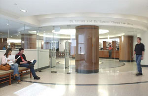 Two students read on a bench outside of the law library. A glass wall separates the students from the library. Another student approaches the others from across the hall.