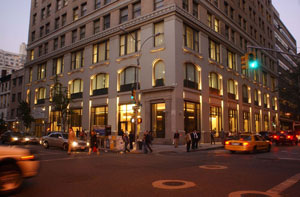 Law school exterior at twilight. A tall building on the corner of Fifth Avenue. Traffic and pedestrians rush by.