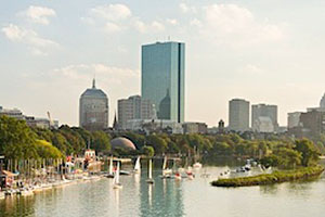 View of Boston. Foreground includes sailboats in a bay. Background includes a blue skyscraper and other tall, gray buildings. A wooded area separates the bay and the city.