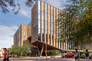 Street view of a brown, angular building striped with vertical rows of windows. Short, green deciduous trees line the front of the building. A red pick-up truck is parked outside. Three diverse women are walking away from the building in different directions.