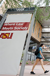 Angled view of an ASU sign that reads: Where Law Meets Society. A woman dressed in athletic wear walks on the sidewalk behind the sign. She is wearing a backpack.