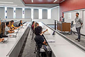 Gray desks are arranged in long rows. Diverse students sit at the desks behind open laptops. Their professor stands at the front of the classroom, next to his small, brown desk.