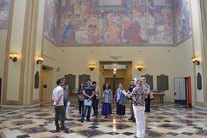 A group of roughly 10 students stand, listening to a lecture in a large, open room. The floors are covered in marble tile, and colorful murals stretch across the upper halves of the high walls.