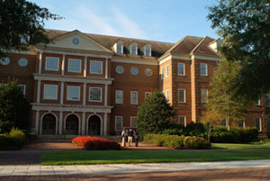 Law school exterior. The building is brick with white accents. Pine trees and shrubs are in front of the building. Deciduous trees are in view.