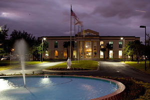 The sky is dark and purple. In the foreground, a blue fountain sprays water into the air. A white statue stands in front of a series of flying flags that lead up to the law school entrance.