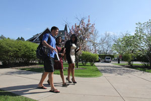 Three diverse students walk along a broad footpath, chatting. Shrubs and grass surround the path. A Victorian-style building peeks out from behind the students, in the background.