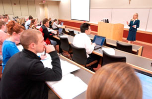 Students listen to their professor give a lecture. Some students have laptops opened in front of them; others, notepads and paper.