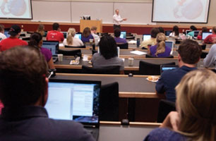 Students listen to a professor give a lecture. The room layout is the same as in the previous photo; however, the professor is different.