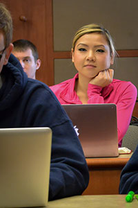 A female student rests her head on her fist while listening to a lecture. A laptop is open in front of her.