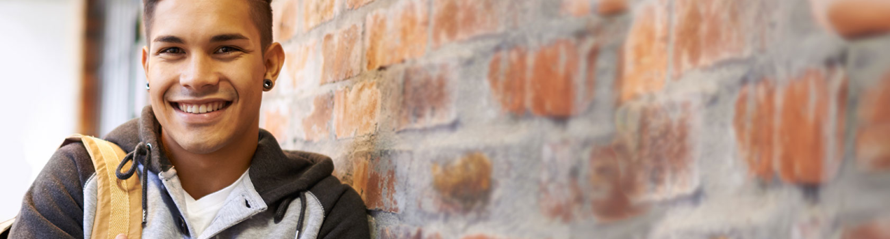 Student with backpack over shoulder, leaning against brick wall.