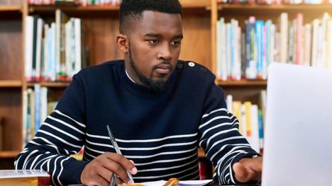 Law student taking notes and looking at laptop in the library.