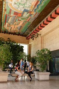 Students chat on benches set between small, potted trees in a foyer. A colorful mural spreads across the ceiling above the students.