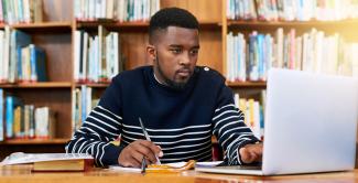 Law student in library, taking notes, and looking at laptop.