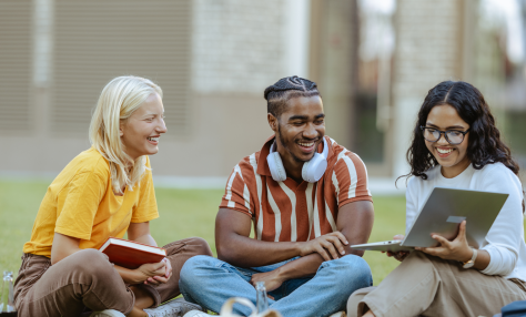 Three students sitting together on the lawn in a campus setting.