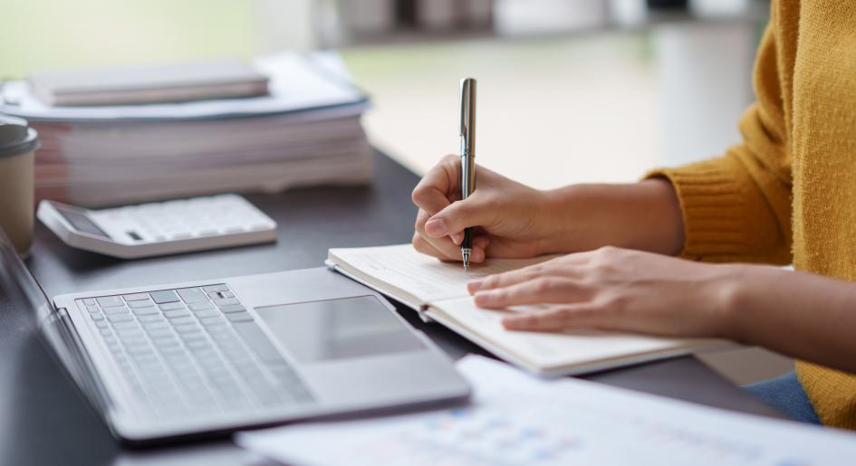 Someone writes in a notebook in front of a computer.