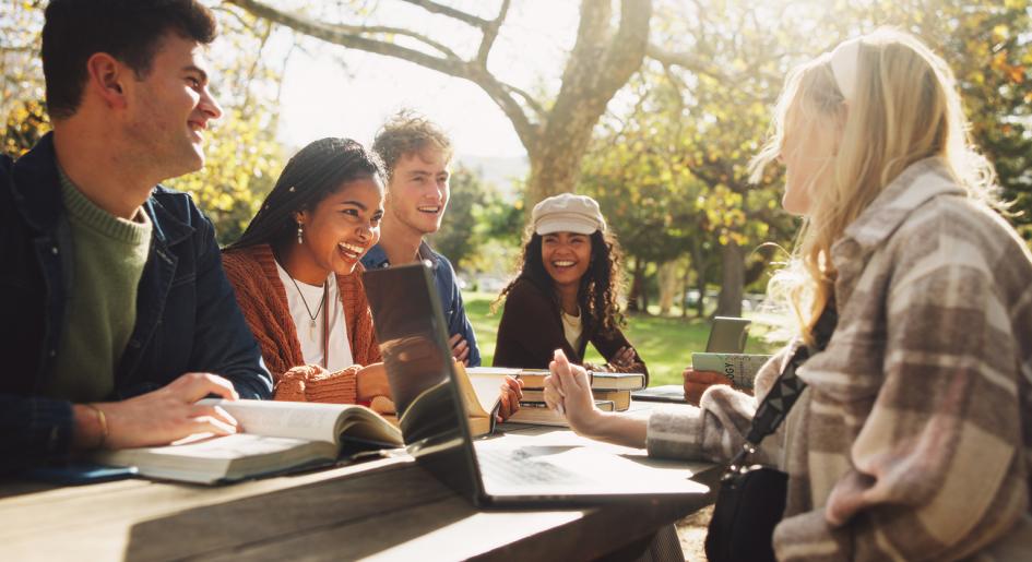 Group of people conversing outside at picnic table with laptops and books.