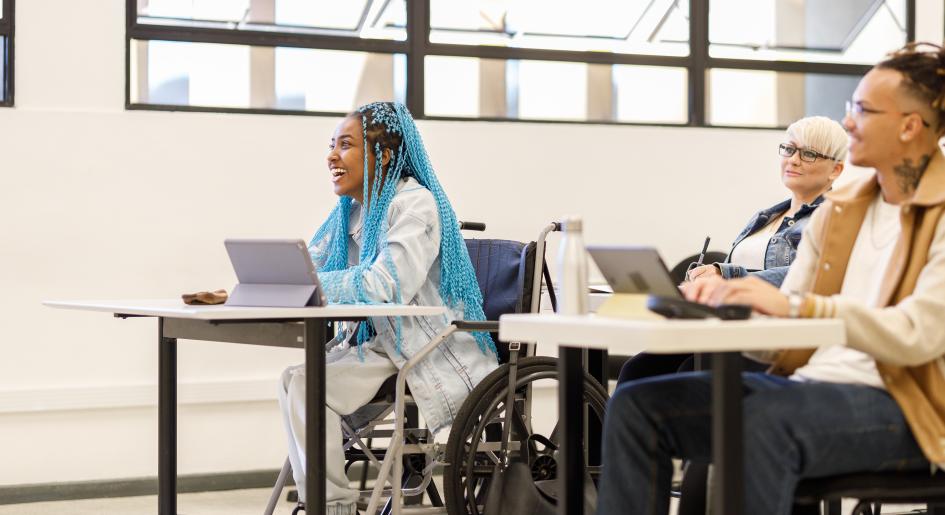 A woman with blue hair sits in a wheelchair in a classroom of students