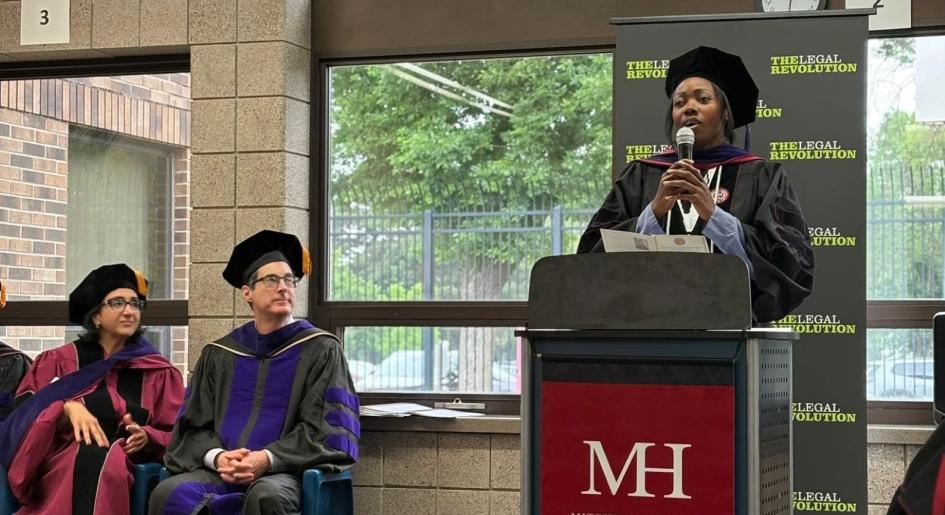 A woman wearing a graduation robe speaks at a podium