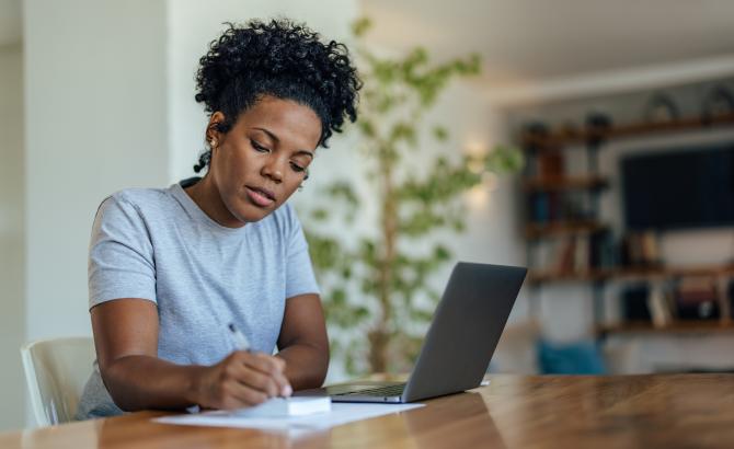 A woman writes on a notepad next to a laptop