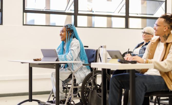 A group of students sit in a classroom. In the center is a wheelchair user