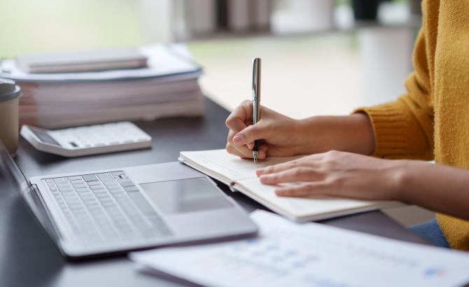 Someone writes in a notebook in front of a computer.